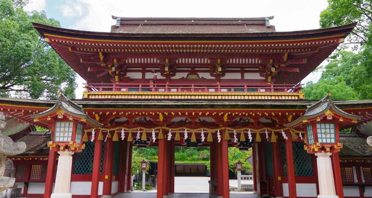 The vividly painted red and gold Romon gate of a Shinto shrine stands symmetrically framed by lanterns and lush greenery.