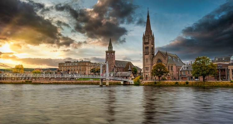 Riverside scene in Inverness with stone church spires and bridge lit by a vibrant sunset sky.