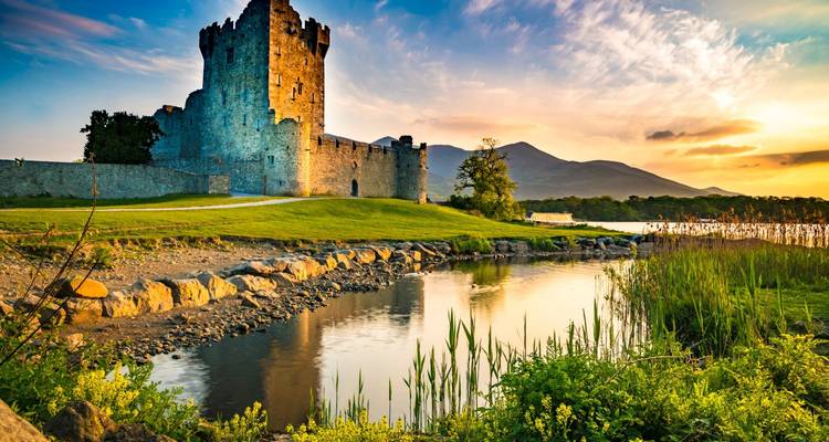 Medieval stone castle by a reflective lake with mountains in the background during sunset light.