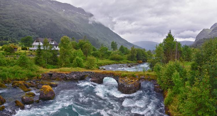 Wild Norwegian valley with rushing river under stone arch bridge, surrounded by green forested mountains on a misty day.