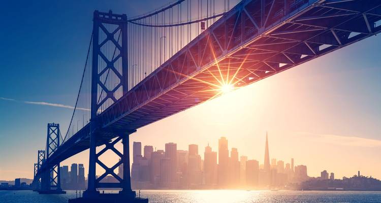 Dramatic sunrise light bursts through the Bay Bridge structure with San Francisco’s skyline softly back-lit.