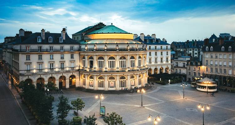 La ópera circular de Rennes brilla con calidez sobre una plaza adoquinada vacía al anochecer.