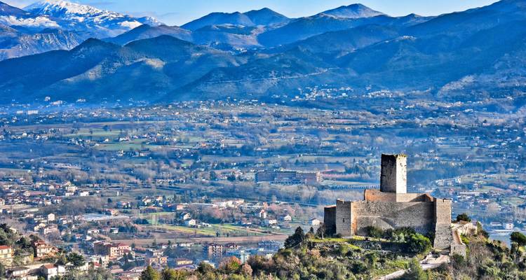 Castillo de piedra encaramado en una colina que domina un amplio valle con montañas nevadas a lo lejos.