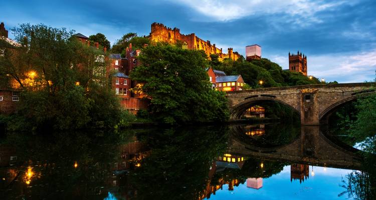 Durham Castle and Cathedral illuminated at dusk above the River Wear and stone bridge reflections.