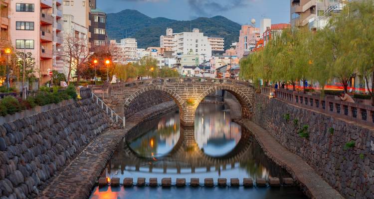 Historic double-arch stone bridge reflected perfectly in a narrow canal flanked by city buildings and hills.