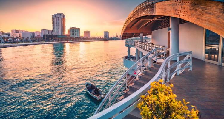 Sunset colors over a seaside promenade with modern buildings and a small fishing boat below.