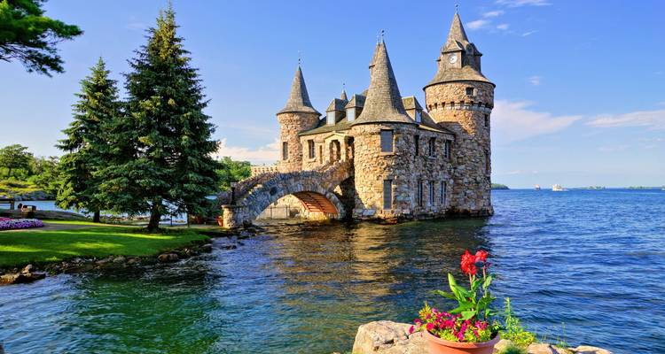 Stone Boldt Castle with pointed turrets rising from an island in a blue river, flowers in foreground.