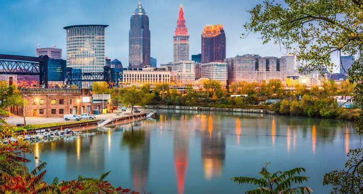 Die Skyline von Cleveland spiegelt sich im Cuyahoga River während der frühen Abendstunden wider, wobei farbige Lichter zu leuchten beginnen.