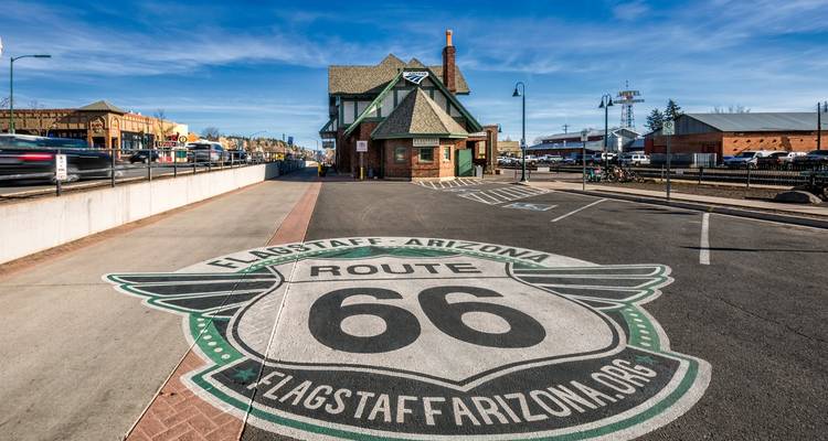 Historic Route 66 Schild auf dem Pflaster in Flagstaff gemalt mit Vintage-Bahnhof unter strahlend blauem Himmel.