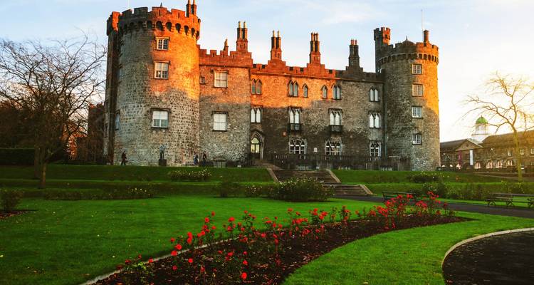 Sunlit stone Kilkenny Castle with blooming rose garden in foreground.