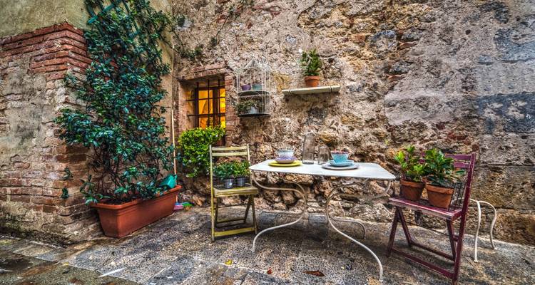 Charmante cour rustique avec table vintage, chaises colorées et plantes en pot contre un mur en pierre.