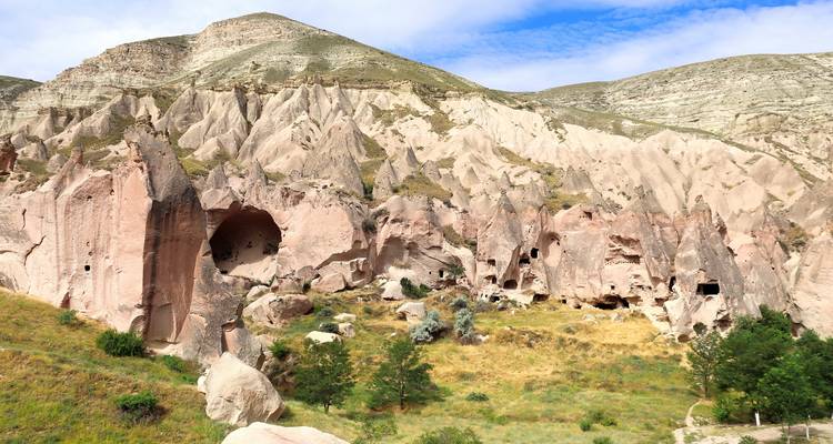 Jagged rock formations and cave dwellings set in rolling Cappadocian landscape under patchy blue sky.