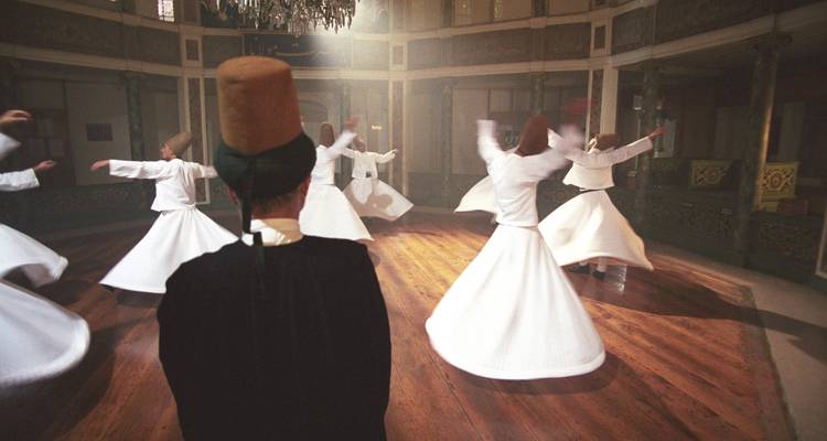 Whirling dervishes in white robes spin inside an old wooden-floored hall while a figure in a tall felt hat looks on.