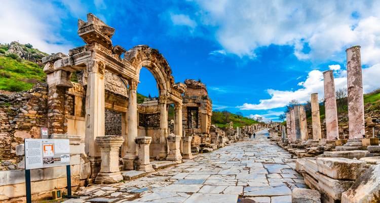 The marble colonnades and arch of the Curetes Street in Ephesus line a sunlit Roman road beneath a bright blue sky.