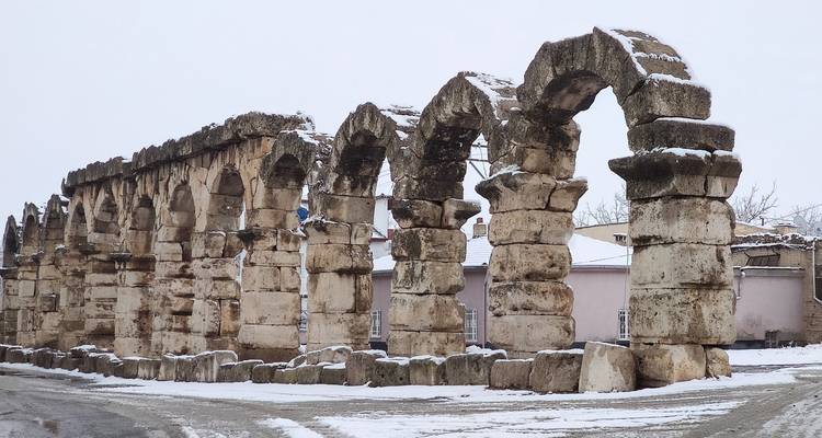 Snow dusts the weathered stone arches of a ruined aqueduct standing in a quiet wintry townscape.