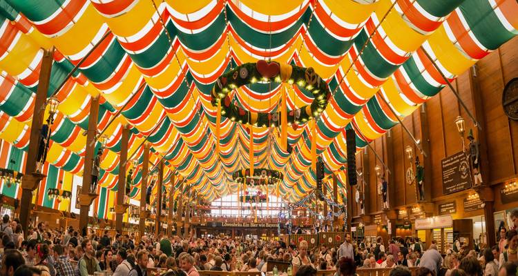 Das farbenfrohe gestreifte Zeltdach eines Oktoberfest-Bierzelts bedeckt Menschenmassen feiernder Gäste, die an langen Gemeinschaftstischen in München sitzen.