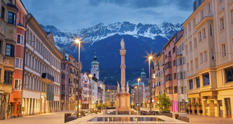 Abendliche Lichter entlang der Maria-Theresien-Strasse in Innsbruck mit schneebedeckten Alpen, die sich dramatisch hinter den farbenfrohen Bürgerhäusern erheben.
