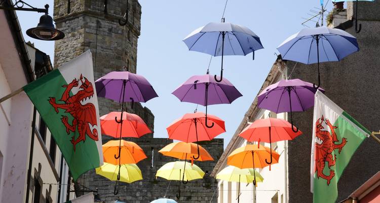 Des parapluies colorés suspendus au-dessus d'une ruelle galloise étroite avec des drapeaux à dragon et une tour en pierre.