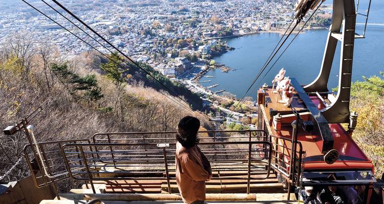 Ein einsamer Besucher blickt von einer Seilbahnplattform hoch über dem Kawaguchiko-See auf die panoramische Aussicht auf die Stadt und das Wasser darunter.