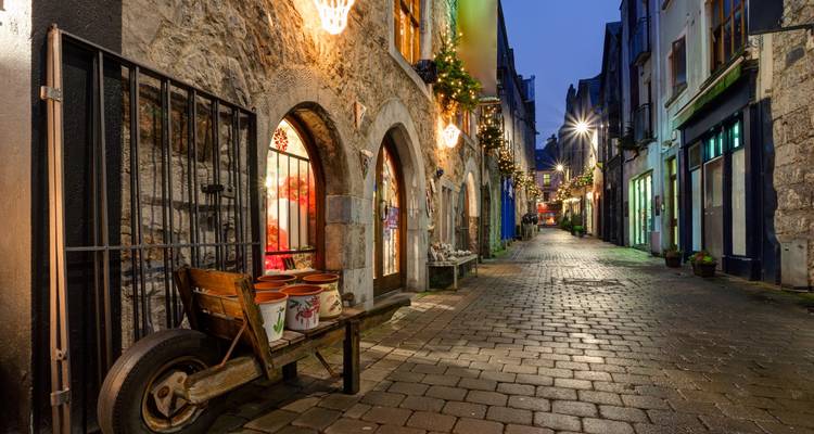 Cobblestone pedestrian lane lined with stone shopfronts and warm lights in Galway at dusk.