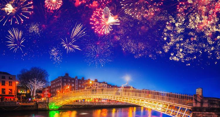 Fireworks burst over Dublin’s Ha’penny Bridge and colorful riverside buildings reflected in the water.