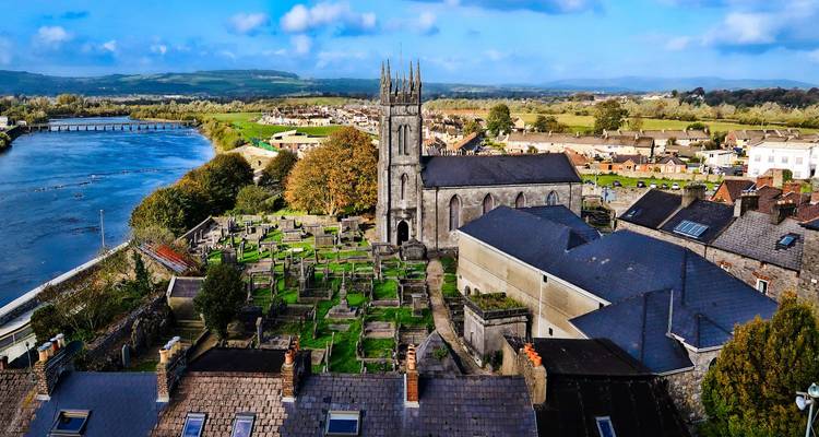 Aerial view of a riverside Irish town with stone church and old cemetery surrounded by green countryside.