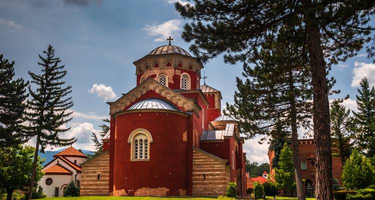 Red-walled Žiča monastery surrounded by tall pines under a partly cloudy sky.