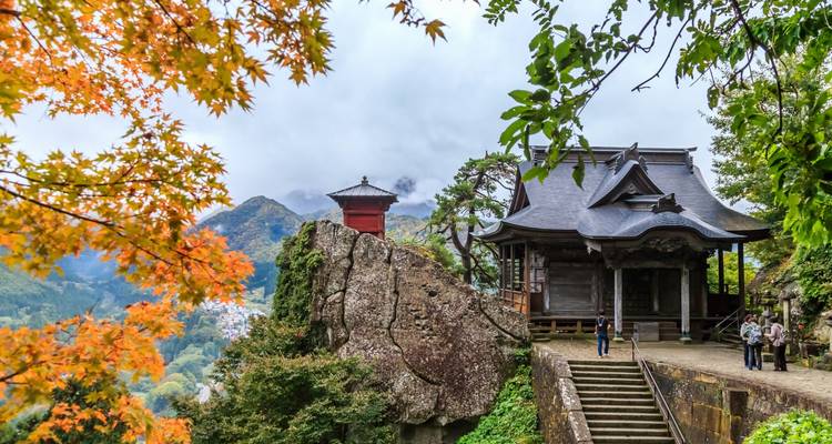 Salle de temple en flanc de montagne à Yamadera encadrée par des feuilles d'érable d'automne colorées avec des pics brumeux au-delà.