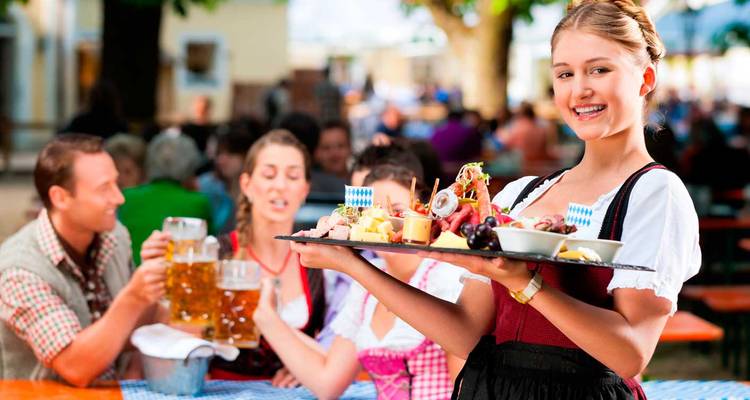 Serveuse souriante en dirndl traditionnel servant des collations bavaroises et de la bière dans un jardin à bière en plein air.