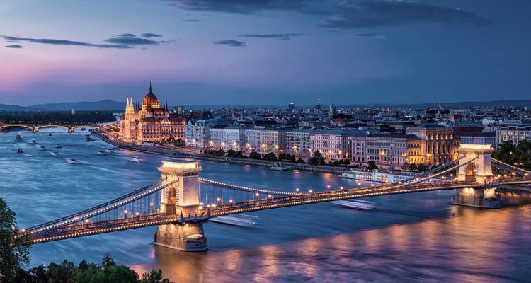 Evening panorama of an illuminated suspension bridge crossing a wide river toward a grand parliament building.