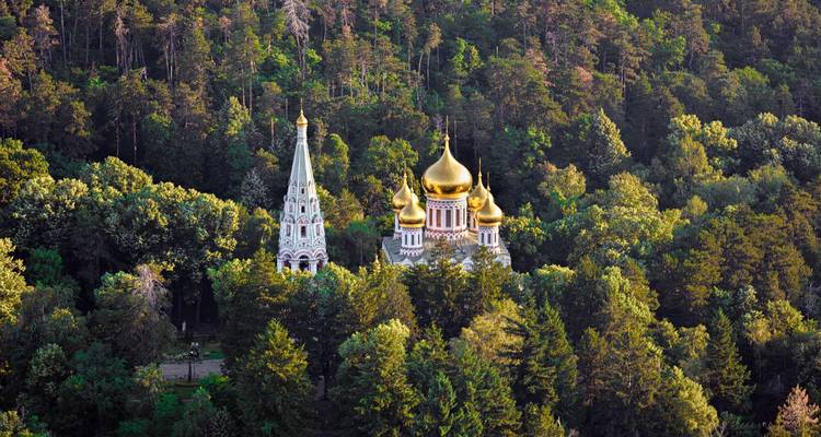 Golden-domed Shipka Memorial Church nestled in a dense green forest viewed from above.