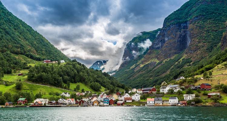 Small village of colorful houses nestles between steep, lush fjord walls under dramatic clouds.