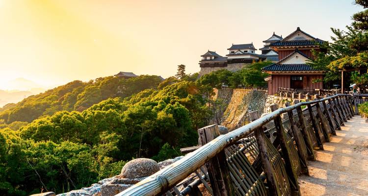 El Castillo Matsuyama bañado por el sol en la cima de una colina domina la exuberante vegetación y las montañas lejanas durante la hora dorada.