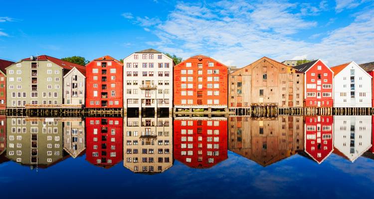 Row of colorful wooden warehouses perfectly reflected in a glassy river beneath a blue sky.