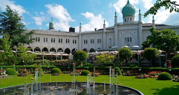Ornate palace building with domes overlooks manicured gardens and fountains on a sunny day.