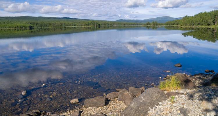 Clear lake reflects scattered clouds and surrounding forested hills with rocky shoreline in foreground.
