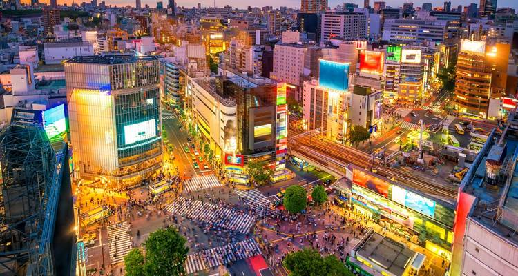 Vue aérienne dynamique du carrefour de Shibuya rempli de foules, d'enseignes lumineuses et de trains filant à travers la soirée tokyoïte.