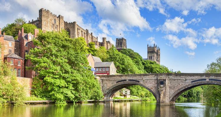 Durham Castle and Cathedral overlook a stone bridge spanning a calm, tree-lined river.