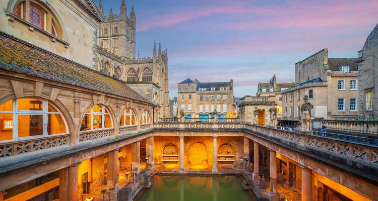 Historic Roman Baths complex glowing at dusk with Bath Abbey towers rising behind.