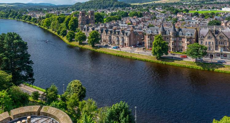 Riverside panorama of Inverness with stone buildings, lush trees and rolling countryside beyond.