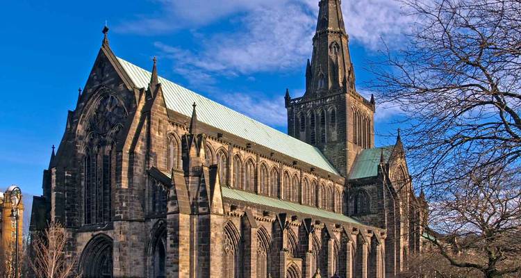 La cathédrale gothique en pierre de Glasgow sous un ciel bleu dégagé avec des branches d'arbres dénudées encadrant la scène.