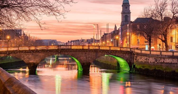 Evening view of a lit stone bridge spanning the River Liffey with the Dublin skyline glowing at sunset.