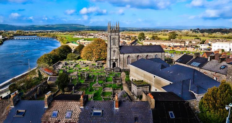 Bird’s-eye view of a church and graveyard beside a wide river in a rural Irish town.