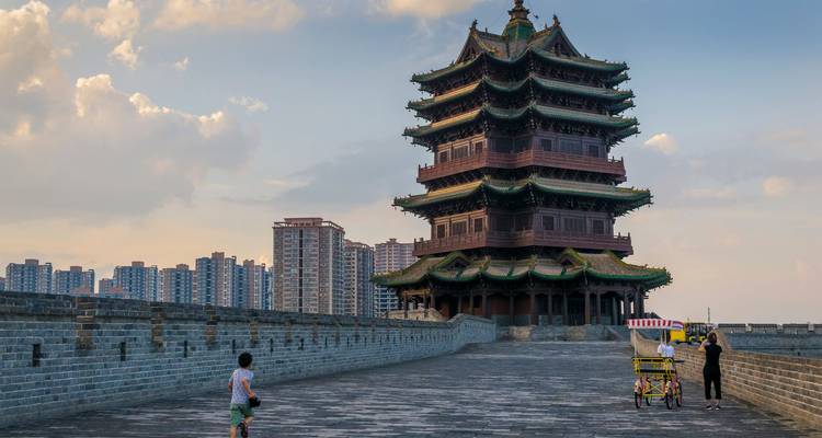 Ancient multi-story wooden pagoda rising above a broad stone city wall with visitors strolling and modern apartment blocks in the background
