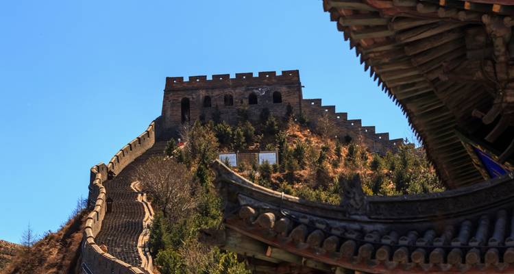 Dramatic upward view of the Great Wall snaking along a steep ridge under a clear blue sky framed by traditional roof eaves