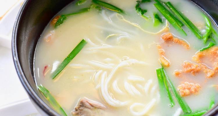 Close-up of a steaming bowl of Chinese noodle soup with green onions and tender meat in a creamy broth