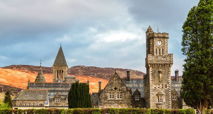 Stone church complex with clock tower set against russet Scottish hills and a moody sky.