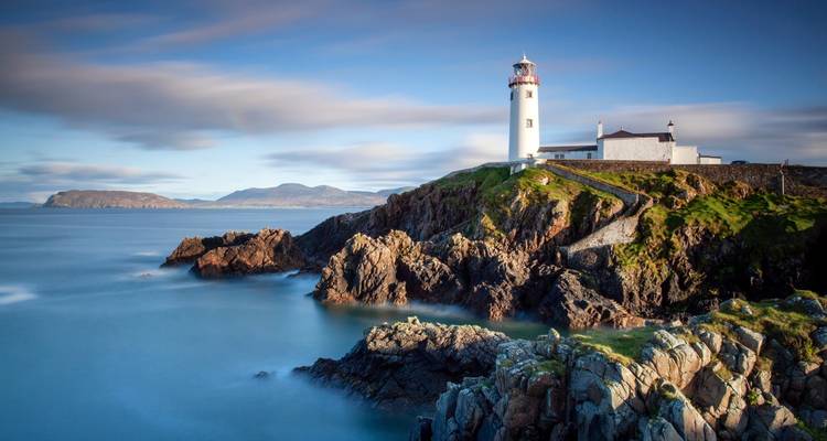 Fanad Head Lighthouse perched on rugged coastal cliffs with silky blue waters and distant mountains.
