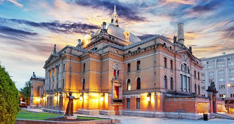 Historic theatre building in Oslo warmly illuminated at twilight with dramatic multi-colored clouds overhead.