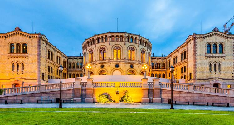 Evening lights illuminate the Stortinget Parliament building in Oslo with a manicured lawn in the foreground.
